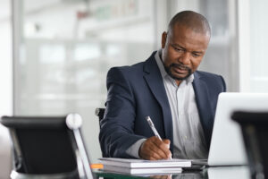 businessman writing notes and using laptop. Mature business man writing his strategy on notebook while using laptop in modern office. Focused black entrepreneur sitting at desk in modern office while working.